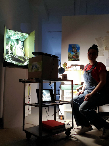 An image of Nicole in their creative environment. They are sitting down next to a desk, surrounded by art equipment and tools. 