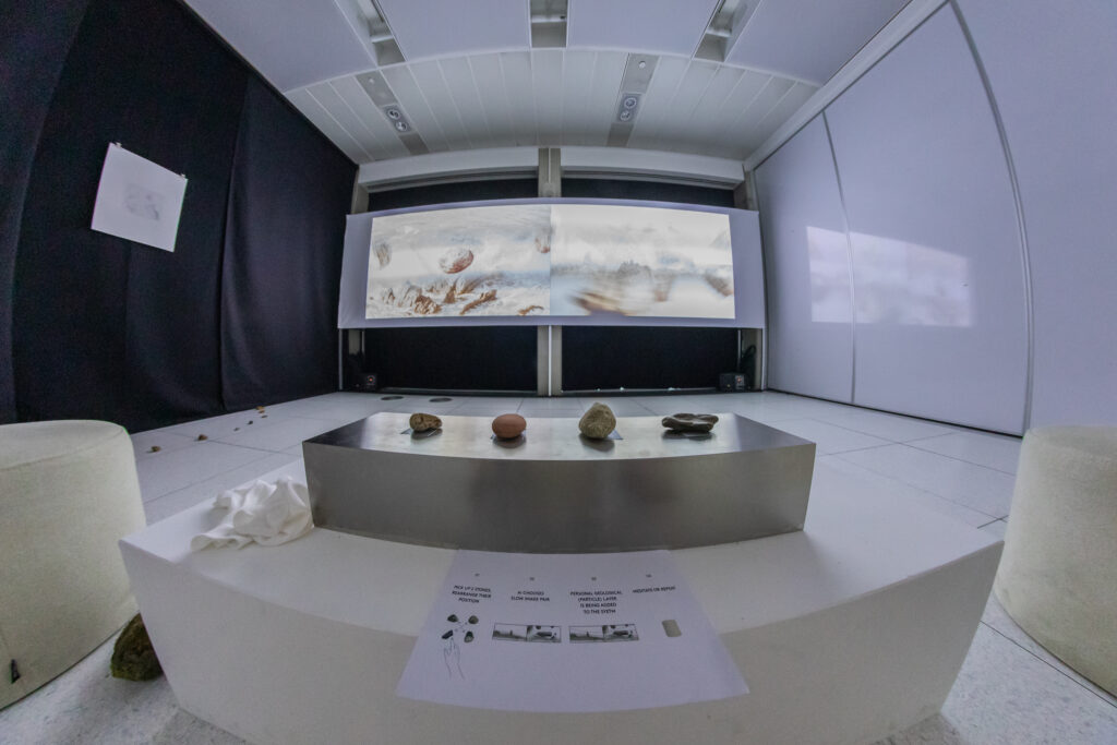 A white exhibition installation with natural rocks/stones set out on one panel, with a background of a white landscape