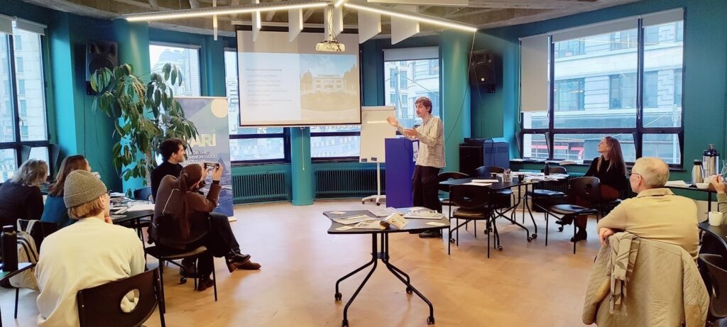 An image taken from the back of a room where individuals are sat around tables watching Ulysse present on a screen. The tables have materials such as papers and images scattered on them.  