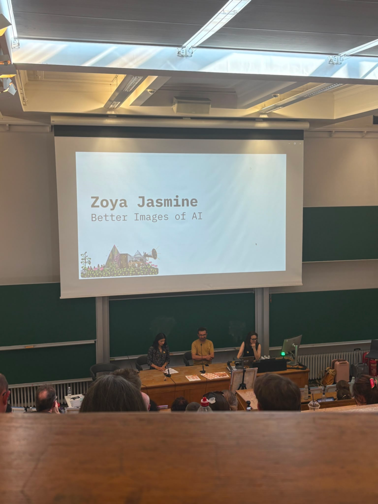 A lecture hall with rows of wooden benches. On the bottom platform is Zoya alongside two other panelists. Above her is a large projection screen with the text, 'Zoya Yasmine, Better Images of AI'.