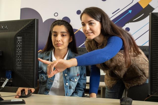 A woman and a man sitting in front of a computer screen, pointing at something on the screen and talking, with a colourful stncil design on the wall behind them.